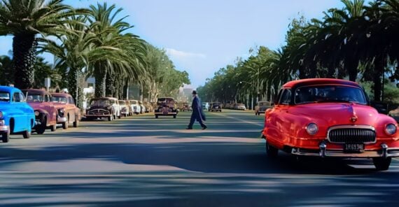 A person crosses a wide, palm tree-lined street filled with vintage cars in various colors, including a prominent red car in the foreground and a blue car to the left, under a clear blue sky.
