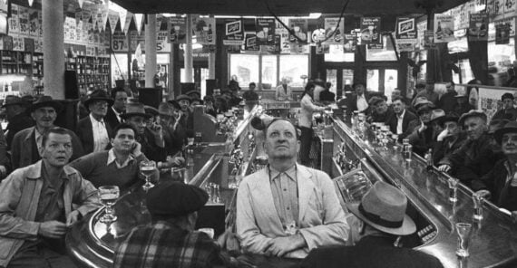 A group of people in a crowded bar watch something off-camera. Most appear focused or tense. Glasses line the bar, and banners hang from the ceiling. The atmosphere suggests anticipation or suspense.