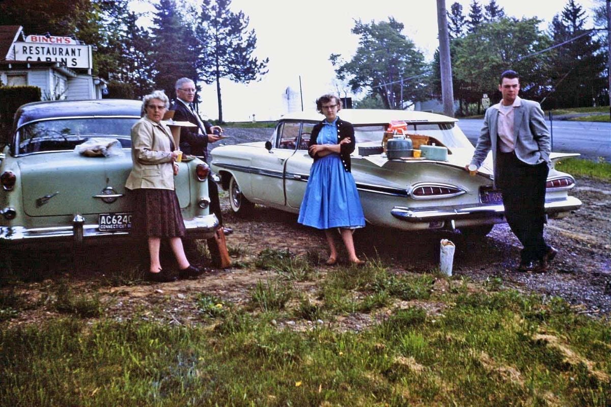 Four people stand beside two vintage cars parked on grass near a roadside, with trees and a restaurant sign in the background. The group appears to be enjoying a roadside meal with food items on a car trunk.