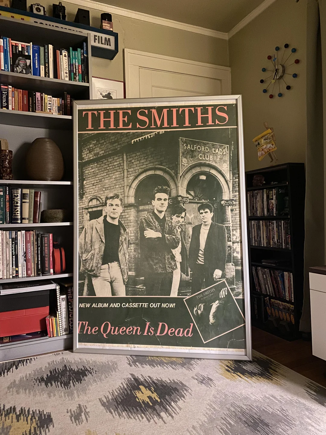 A framed poster of The Smiths' album "The Queen Is Dead" stands in a cozy, book-filled room with shelves, a rug, and a modern clock on the wall.