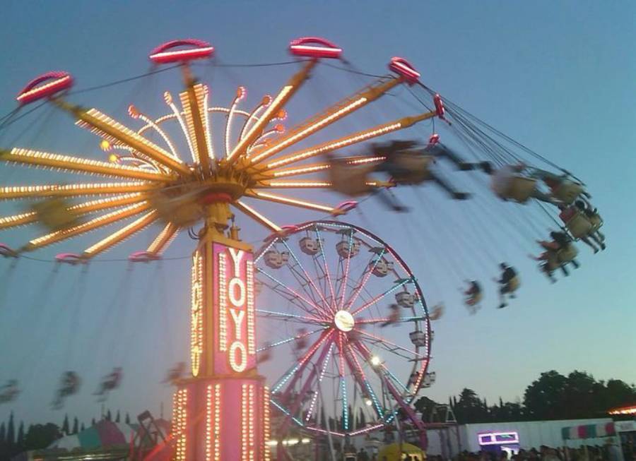 A brightly lit Yo-Yo swing ride spins riders at dusk near a Ferris wheel at a fairground, with blurred motion showing the speed and excitement of the amusement park.