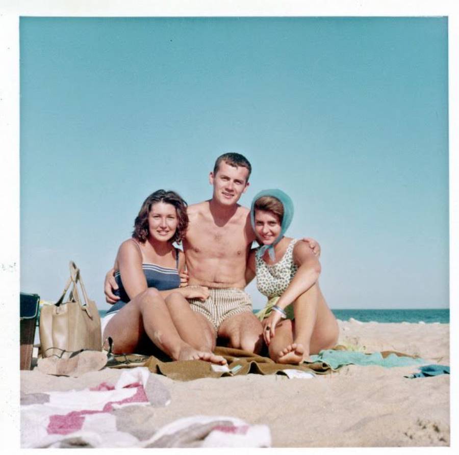 Three people sitting closely together on a sandy beach, smiling at the camera. A woman on each side of a shirtless man; one woman wears a headscarf. The sky is clear and blue. Towels and a bag are on the sand.