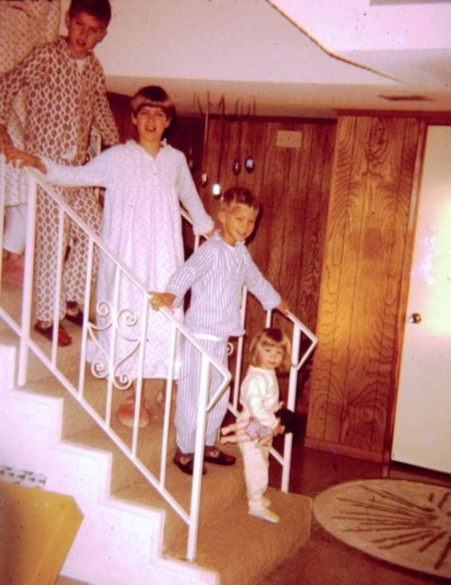 Four children in pajamas stand on carpeted stairs with a white metal railing in a wood-paneled room. Three are older kids on the steps, and a small child stands at the bottom, holding the railing and smiling.