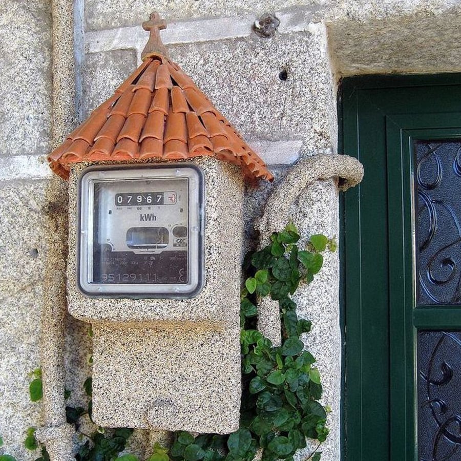 A utility meter installed on a stone wall, covered by a small roof made of red tiles shaped like a miniature house, with some green ivy growing nearby.