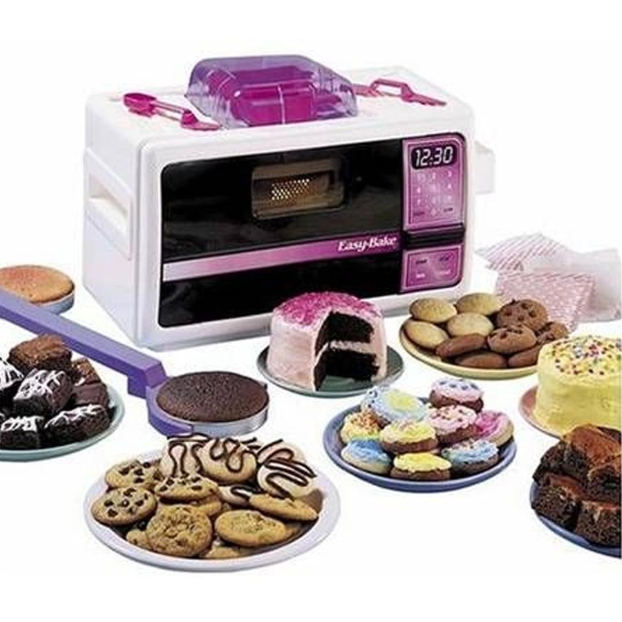A toy Easy-Bake Oven with a digital clock is surrounded by various baked treats, including cookies, cakes, cupcakes, and brownies, all displayed on colorful plates. Baking utensils and paper cupcake liners are nearby.