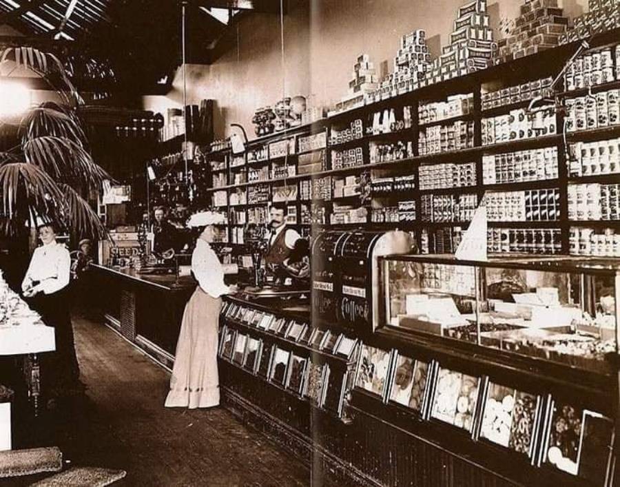 A vintage grocery store with tall shelves filled with canned goods, a glass display case, and two women in early 1900s attire standing at the counter, one being served by a man behind the register.