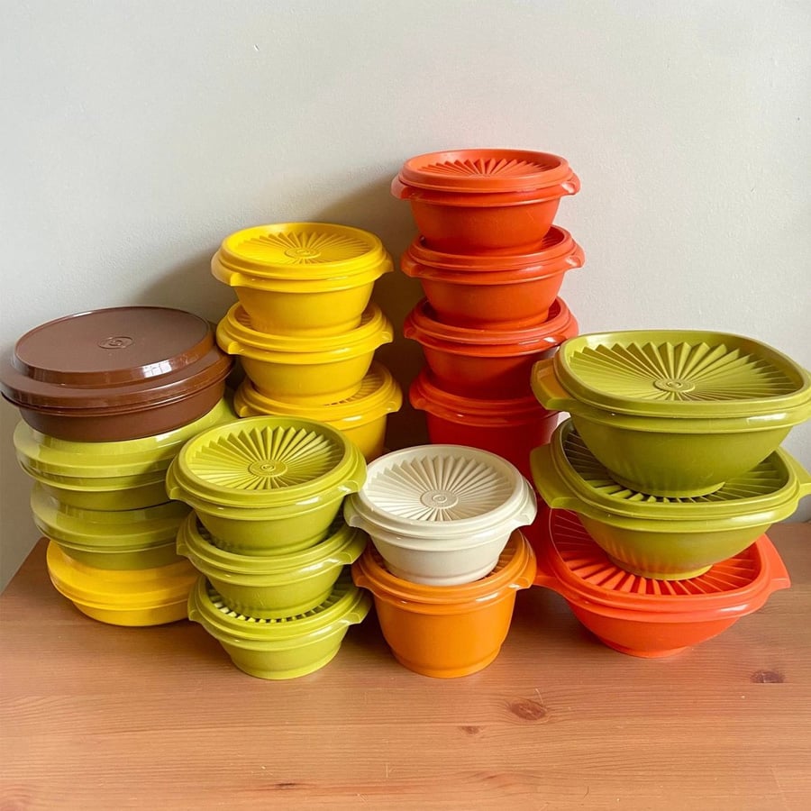A collection of vintage plastic containers in yellow, orange, green, brown, and cream colors, stacked neatly on a wooden surface against a light background.
