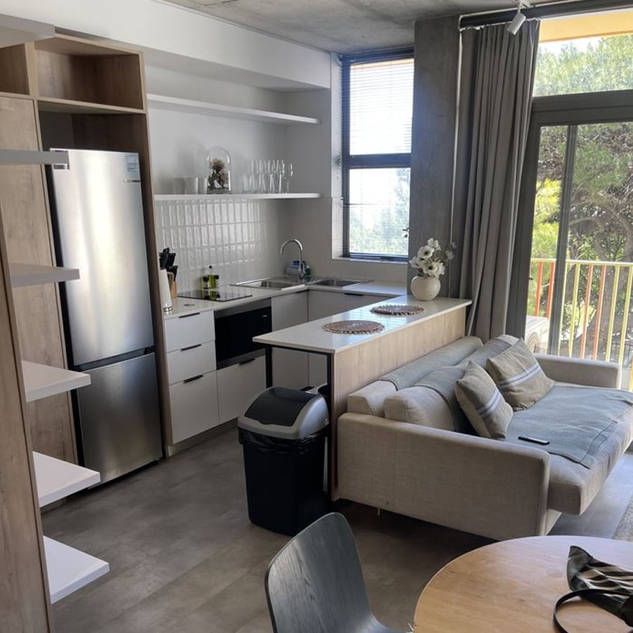 Modern kitchen and living area with light wood cabinets, white countertops, stainless steel fridge, small sofa, dining table, and large window with curtains overlooking greenery.