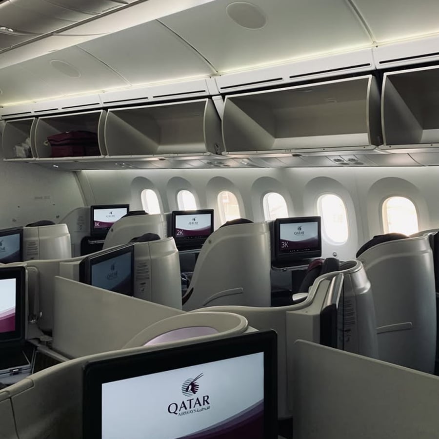Qatar Airways business class cabin with individual seats, personal screens, and overhead bins open. Sunlight comes in through the large windows on one side of the airplane.