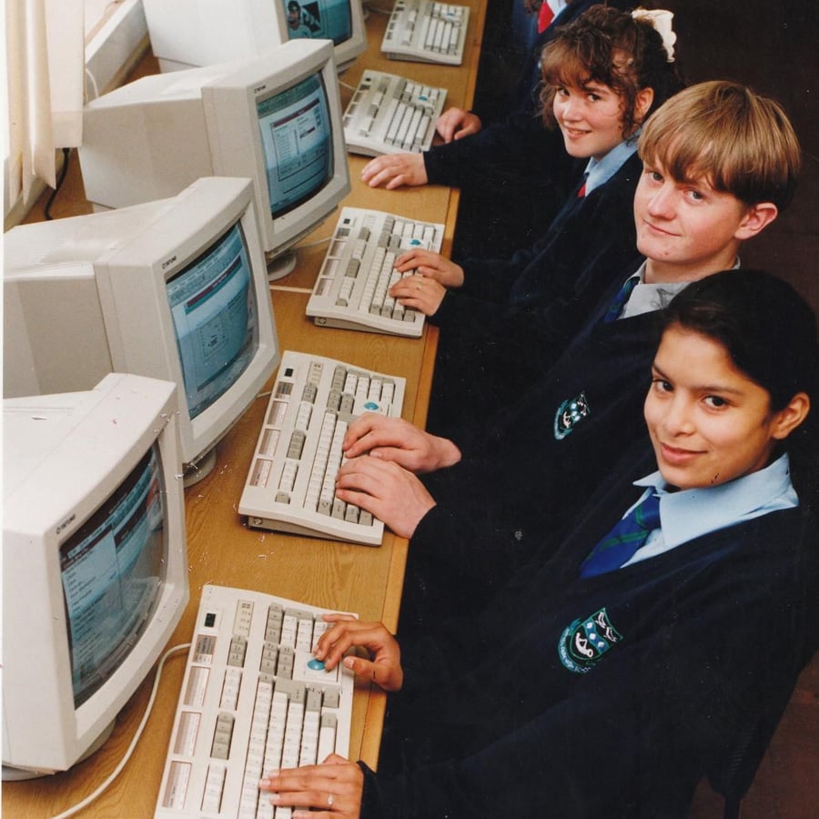 Four students in school uniforms sit at a row of old desktop computers with CRT monitors, typing and smiling at the camera in a classroom setting.