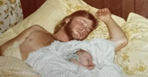 A man with a beard is sleeping on a bed with yellow pillows and floral sheets, cradling a newborn baby under a white blanket beside him. The background shows wood-paneled walls and a floral curtain.