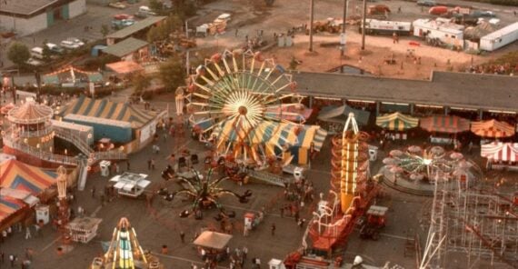 Aerial view of a busy fairground with colorful rides and attractions, including a Ferris wheel, carousel, and tents. Beyond the fair, there is a racetrack and a river, with green hills in the background.