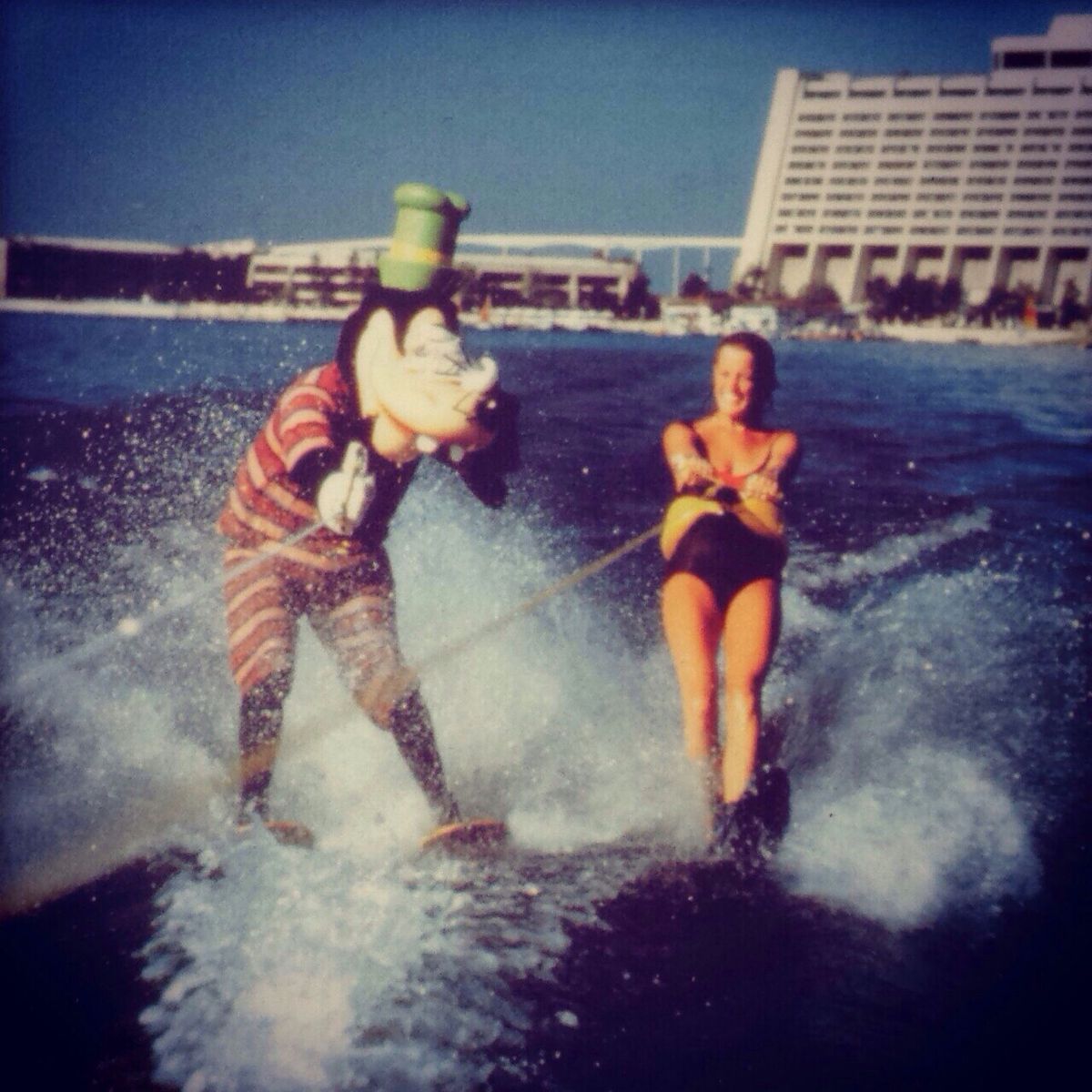 A person in a Goofy costume water-skis next to a woman in a yellow and black swimsuit, both holding onto a tow rope, with a large hotel building and beach in the background under a sunny sky.