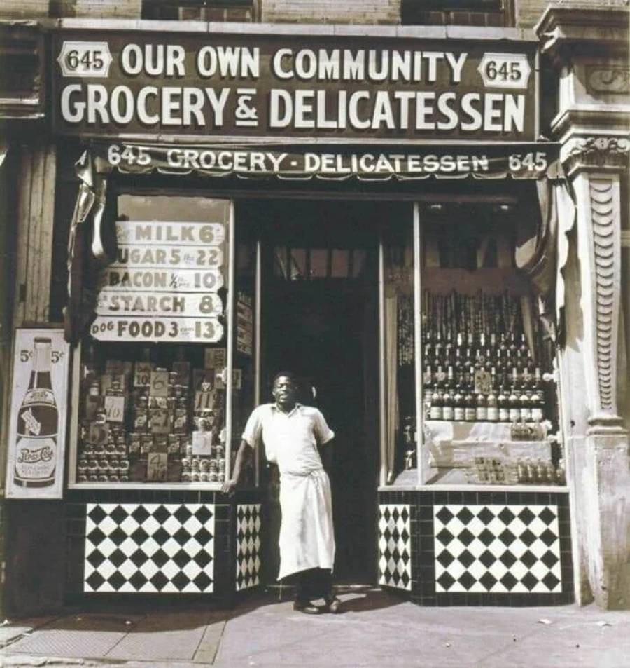 A man in an apron stands in the doorway of a grocery and delicatessen shop with checkered tiles, vintage signs, and shelves of goods in the windows. The shop sign reads “Our Own Community Grocery and Delicatessen.”