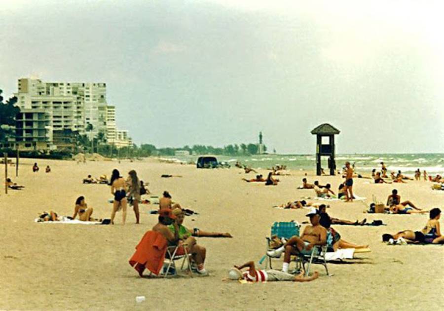 People sunbathe and relax on a sandy beach near the ocean, with some sitting on chairs and others lying on towels. High-rise buildings and a lifeguard hut are visible in the background.