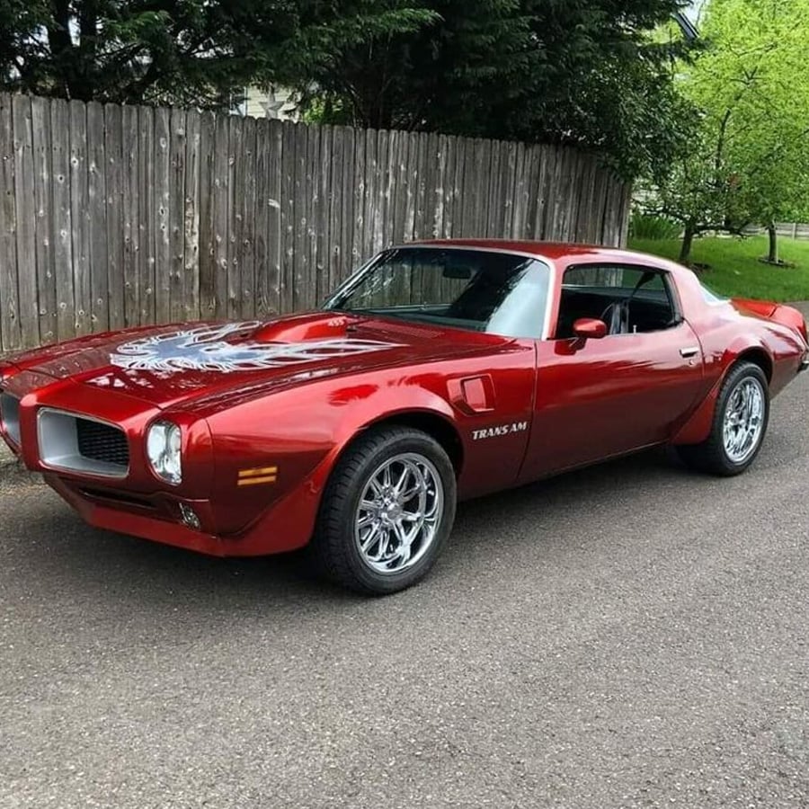 A shiny red classic Pontiac Trans Am car is parked on a road beside a wooden fence and green trees, with a bird decal on the hood.