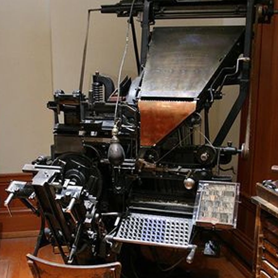 A vintage Linotype machine with a keyboard, metal parts, and a chute for casting type, displayed in a museum setting against a wall.