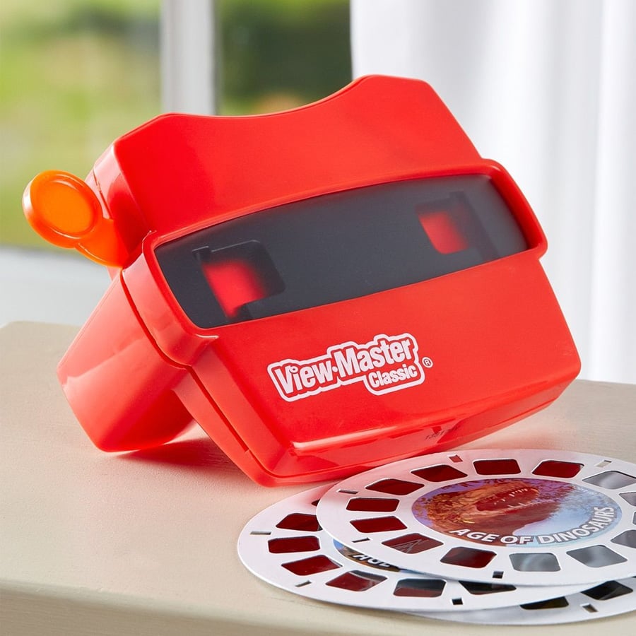 A red View-Master Classic toy sits on a table next to a photo reel labeled “Age of Dinosaurs.” The toy has black lenses and an orange lever on the side.