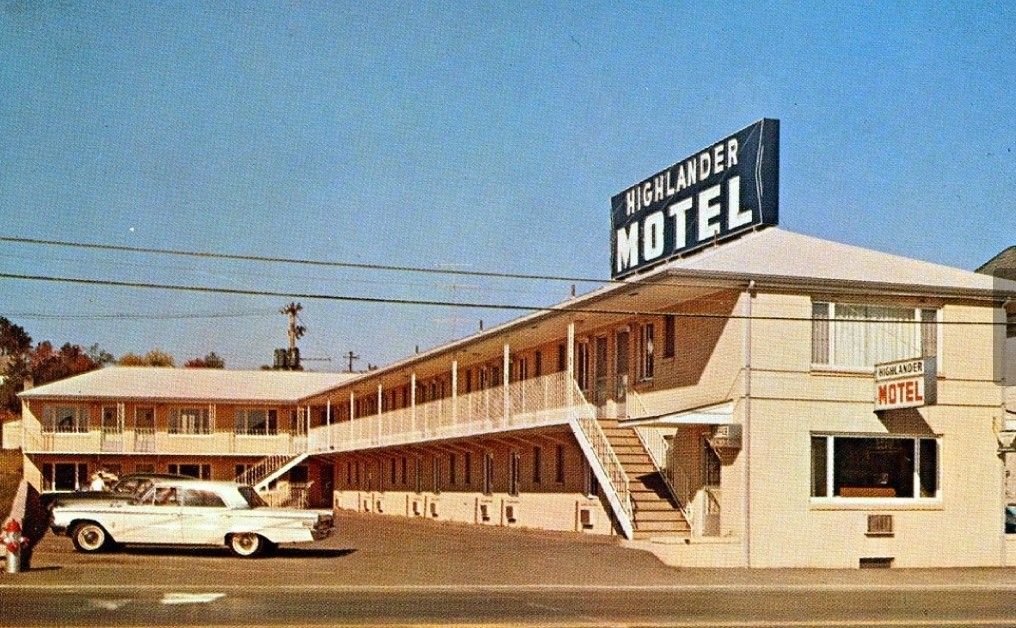 A vintage two-story motel with a large "Highlander Motel" sign on the roof. A classic white car is parked out front, and stairs lead to the second floor. The building has a beige exterior under a clear blue sky.
