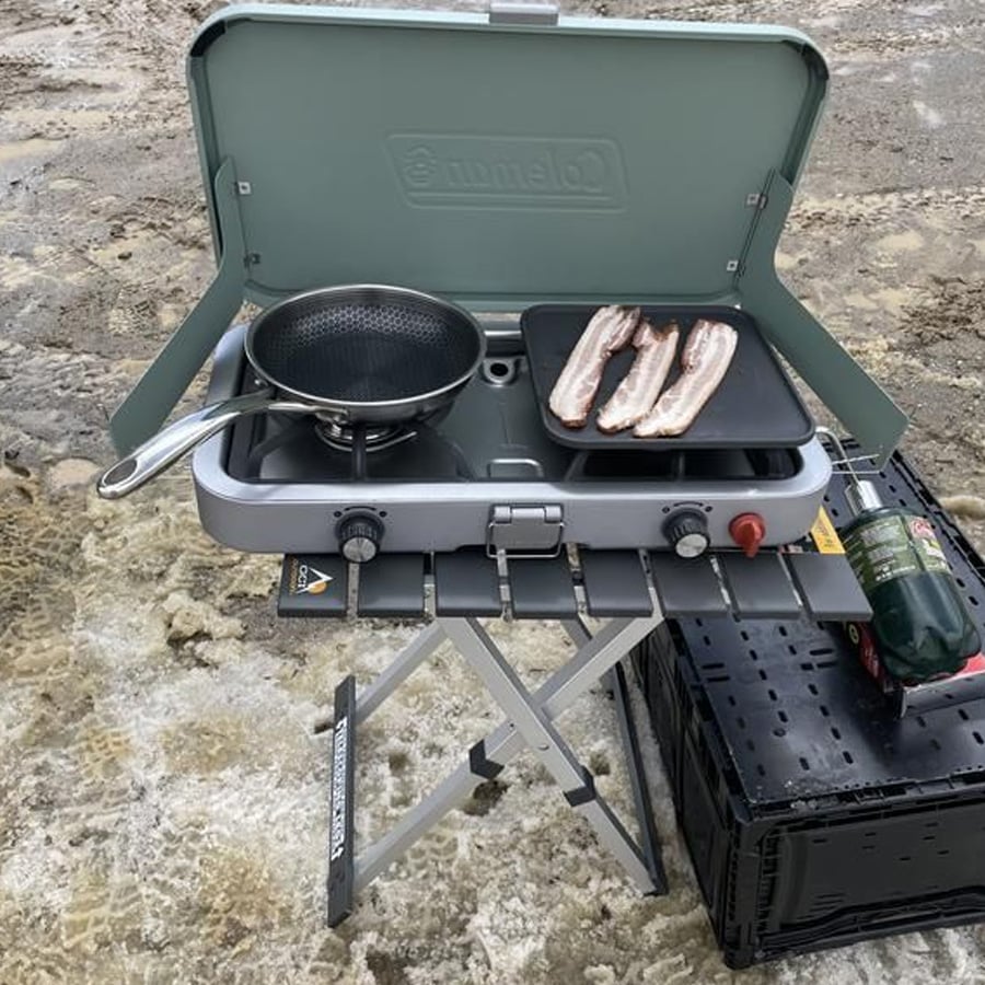 Portable camping stove with a frying pan on one burner and bacon strips cooking on a griddle. The stove sits on a foldable table outdoors on muddy, snowy ground. A green propane tank is attached on the right side.