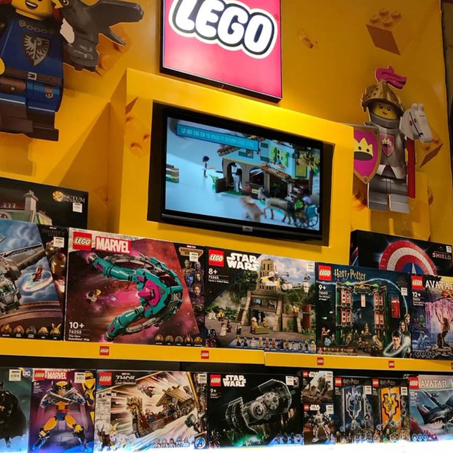 Shelves in a LEGO store display various themed LEGO sets, including Marvel, Star Wars, Harry Potter, and Avatar, beneath a screen showing LEGO animations and a large LEGO logo sign.