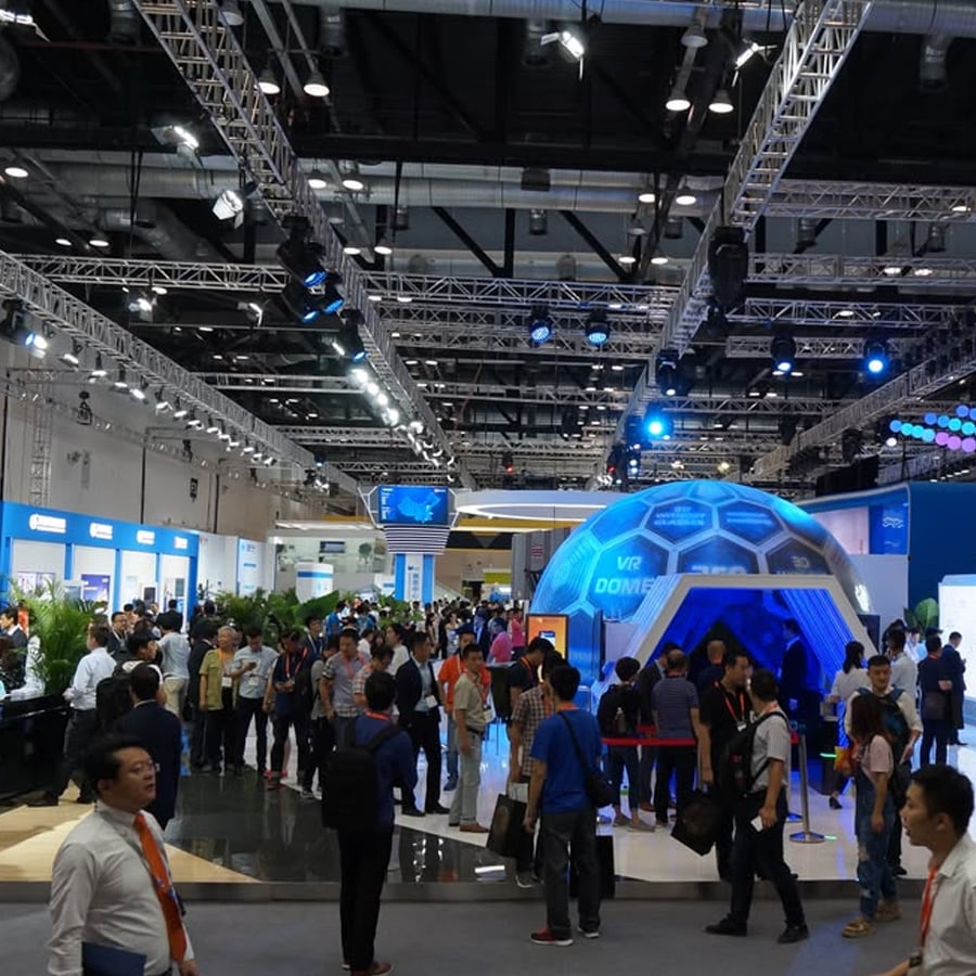 A busy tech exhibition hall with many people gathered around a futuristic, dome-shaped VR booth, bright lights, and various displays in the background.