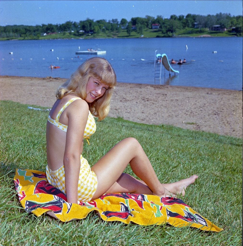 A young woman in a yellow checkered swimsuit sits on a colorful towel on the grass by a sandy lake beach, smiling. The lake, with a slide and boats, and trees are visible in the background under a blue sky.