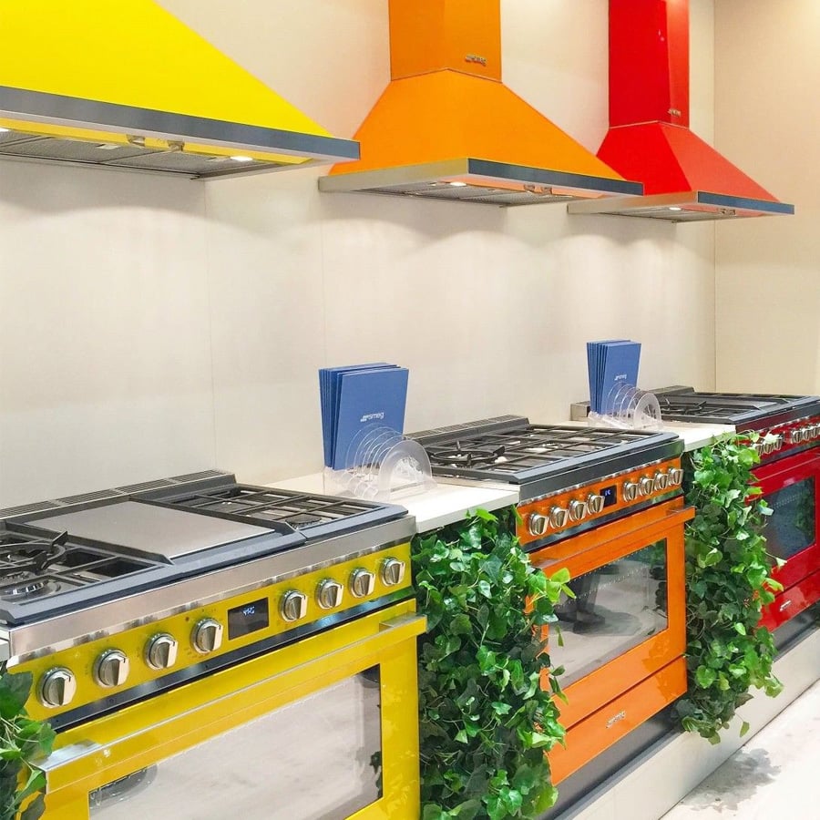 Three stoves with matching yellow, orange, and red colors and hoods are lined up in a modern kitchen, decorated with green plants on the counters.