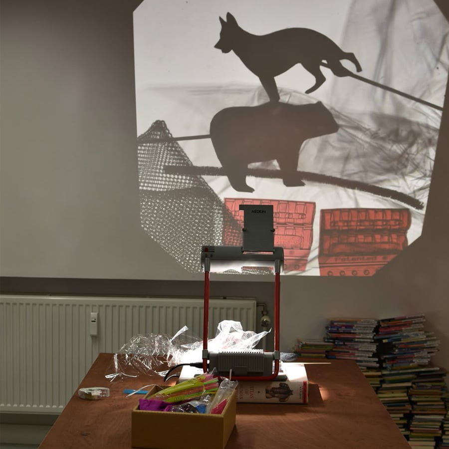 An overhead projector casts animal shadow puppets onto a wall. Supplies and paper are on a table; stacks of books are in the background.