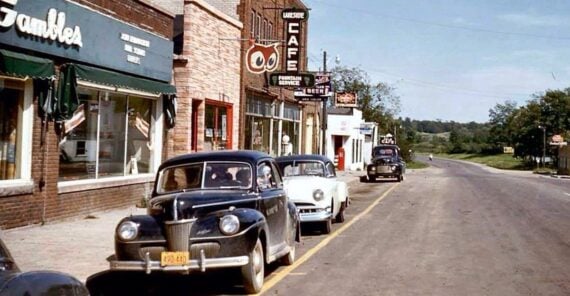 A vintage street scene with classic cars parked along a small-town road, storefronts including a grocery, a cafe, and a Gambles store, under a clear blue sky with trees and open landscape in the background.