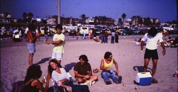 A group of people relaxing on a sandy beach, some sitting on towels or chairs, chatting and enjoying snacks. Cars are parked in the background and palm trees and houses line the horizon under a clear sky.