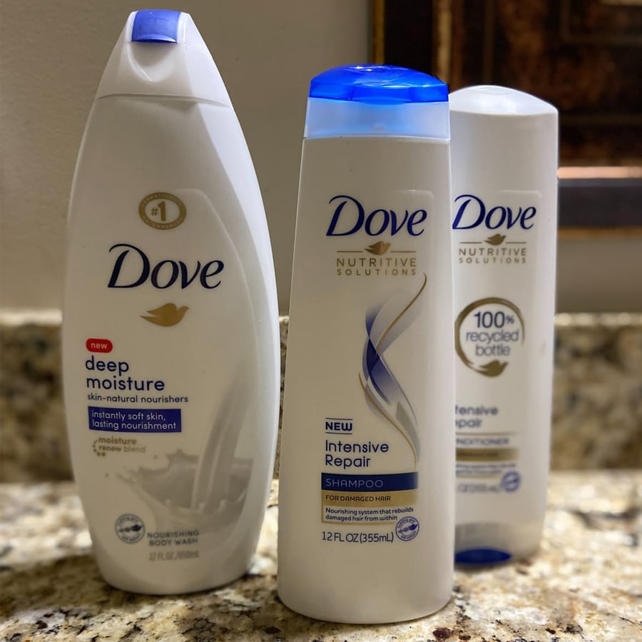 Three Dove bottles on a granite countertop: Deep Moisture body wash, Intensive Repair shampoo, and Intensive Repair conditioner, with a focus on moisturizing and nourishing features.