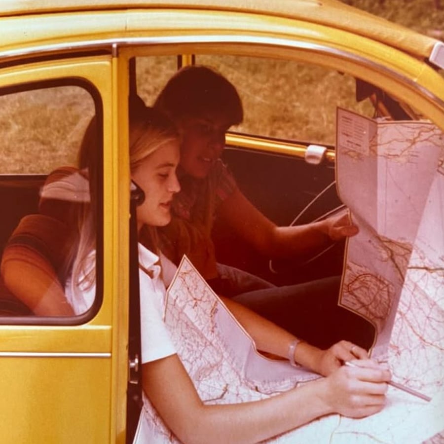 Two young women sit in a yellow car, examining a large paper map together. The car door is open, and they appear focused on planning or navigating their route.