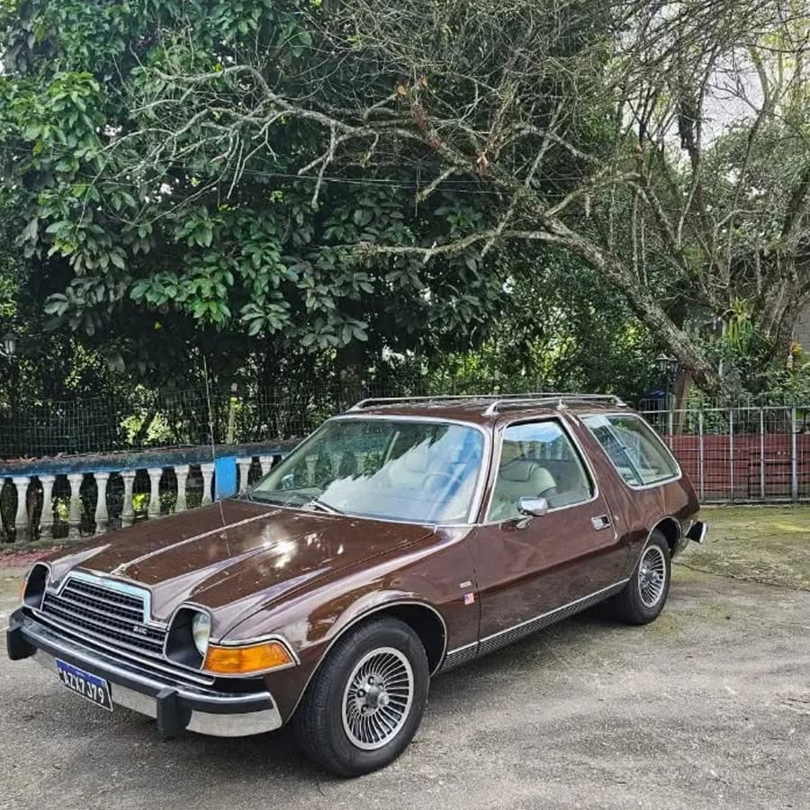 A vintage brown car with a unique rounded design is parked outdoors on a dirt surface, surrounded by trees and greenery.