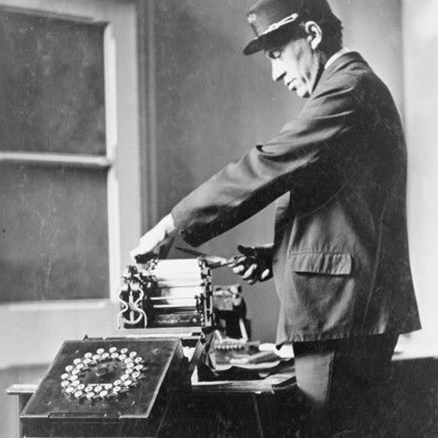 A man in a uniform operates an early typewriter or telegraph machine near a desk with a circular device, in a black-and-white historical photo.