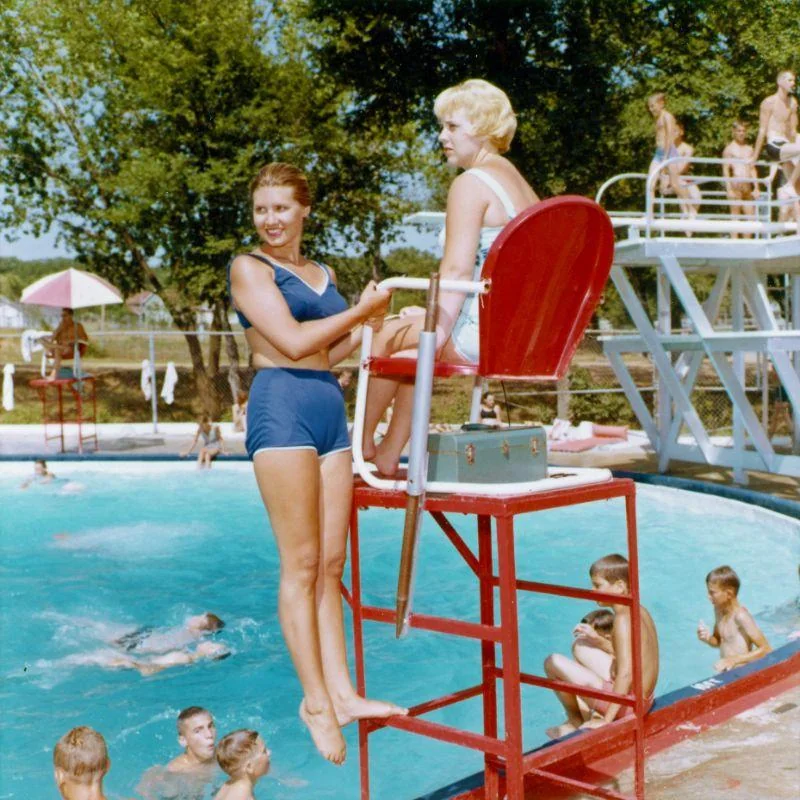 Two women in vintage swimsuits chat at a pool; one stands on the lifeguard chair’s step while the other sits on the chair. Kids swim and play in the background on a sunny day.