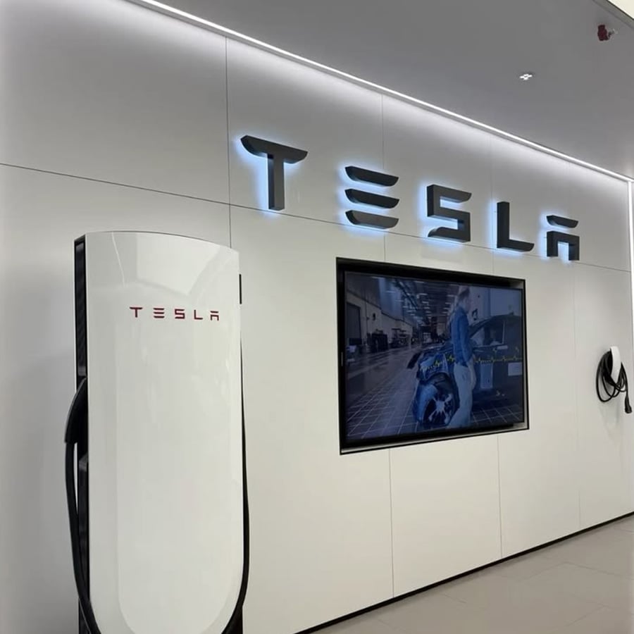 A Tesla showroom features a white charging station, a large screen displaying a car, and the illuminated Tesla logo on a sleek white wall.