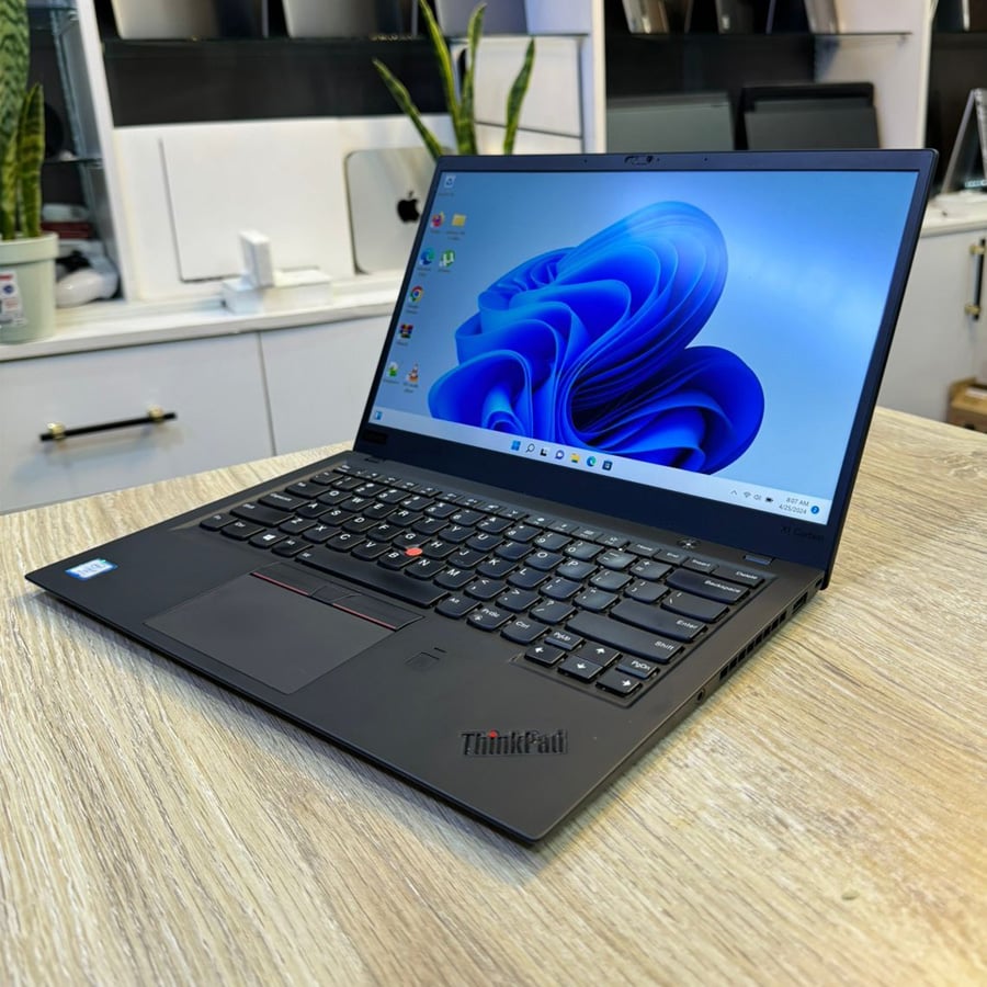 A black Lenovo ThinkPad laptop is open on a wooden desk, displaying the Windows 11 home screen. Office supplies and plants are visible in the blurred background.