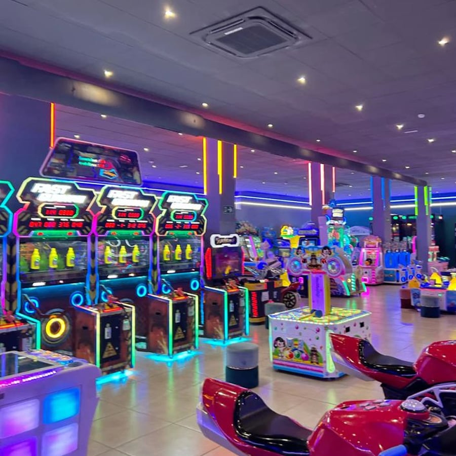A brightly lit arcade with rows of colorful, glowing game machines and racing simulators. The spacious room features neon lights and a shiny tile floor, creating a lively and fun atmosphere.