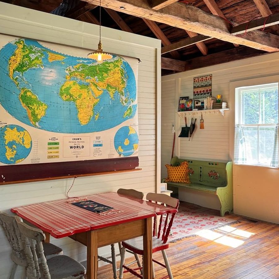 Cozy room with a world map on the wall, a small table with mismatched chairs, a green bench with pillows, wooden floor, exposed beams, and sunlight streaming through a window.