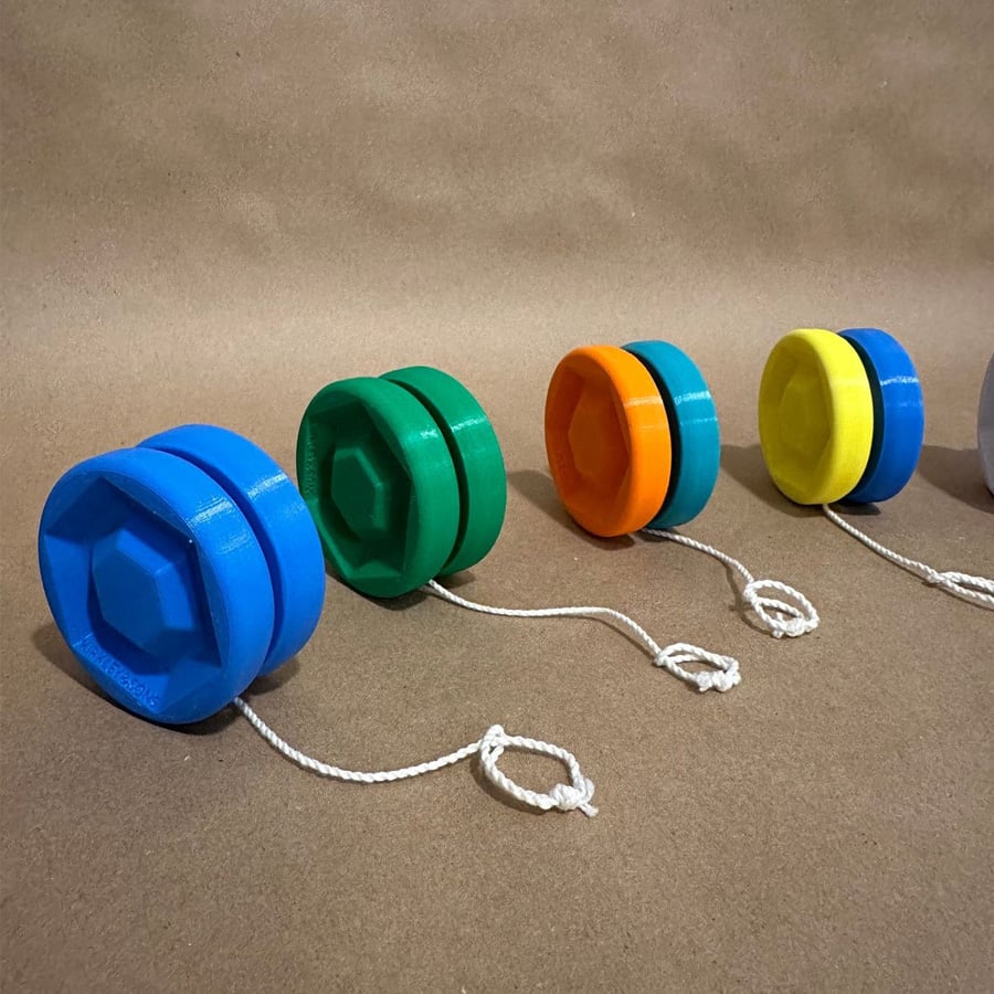 Four plastic yo-yos in blue, green, orange, and yellow are lined up on a brown surface, each with a white string attached, against a plain brown background.
