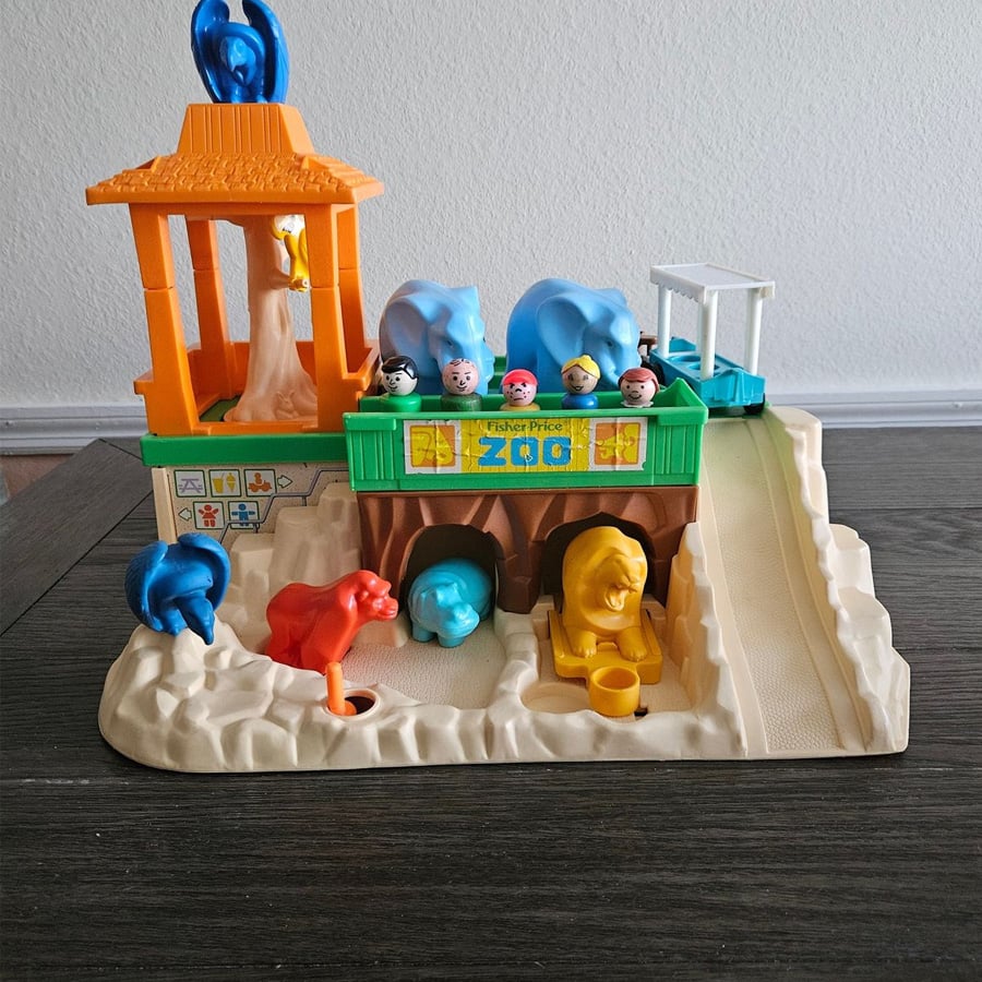 A vintage Fisher-Price zoo playset with animal figures, zookeeper, and children figurines on a brown base, with a slide, animal enclosures, and a yellow feeding station, set on a wooden table.