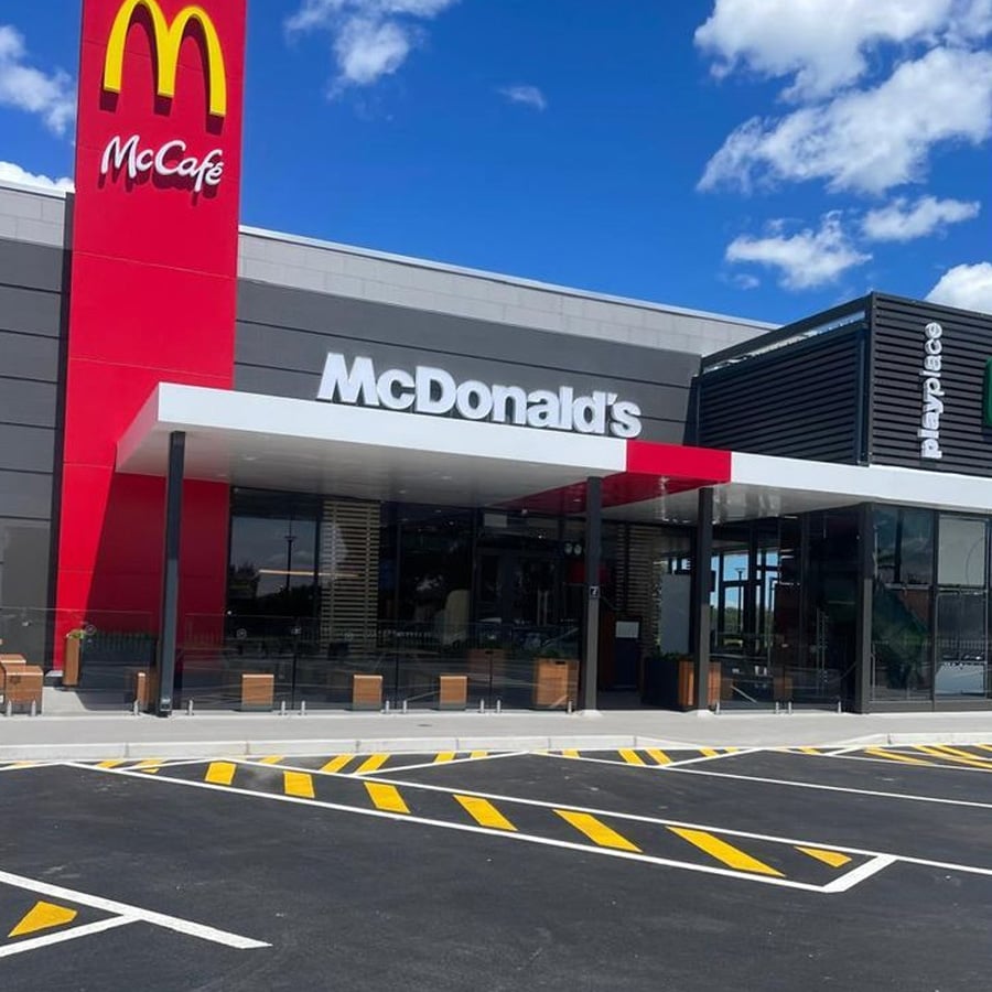 A modern McDonald's restaurant with a McCafé section, outdoor seating, and a playplace area, under a blue sky with scattered clouds. The parking lot in front has yellow striped markings.
