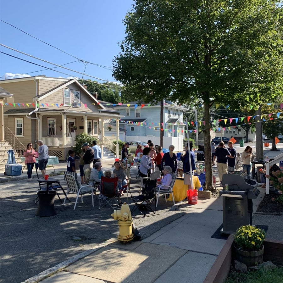A group of people gather on a residential street, socializing among chairs and tables under colorful pennant banners. Trees provide shade and houses line the street. A grill and fire hydrant are visible in the foreground.