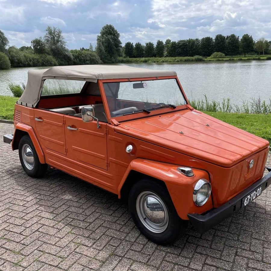 An orange vintage convertible car with a beige soft top is parked on a brick road near a lake, with trees and cloudy sky in the background.