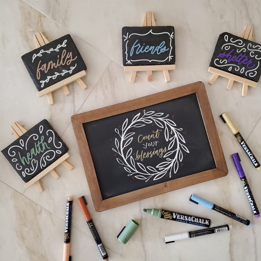 Five mini chalkboards with positive words—“family,” “friends,” “health,” “smile,” and “count your blessings”—are displayed on a floor with colored chalk markers scattered around them.