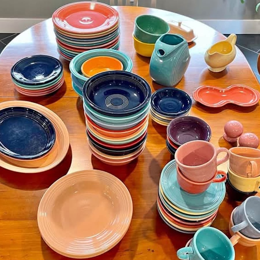 Colorful ceramic dishes, bowls, cups, pitchers, and salt shakers are neatly stacked on a wooden table. The dishware features bright shades of blue, orange, yellow, green, pink, and more.