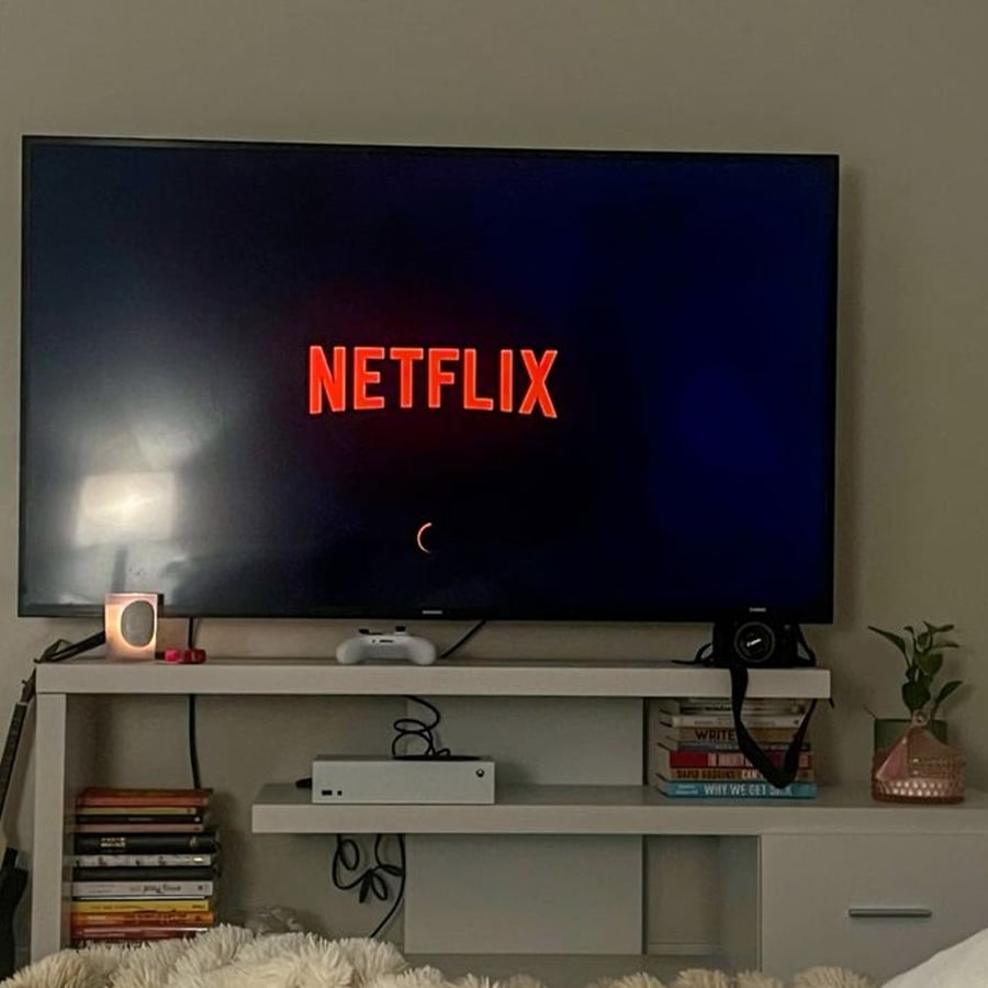 A TV displaying the Netflix logo sits on a white stand, surrounded by a gaming controller, books, a camera, a small plant, and cozy bedding in the foreground.