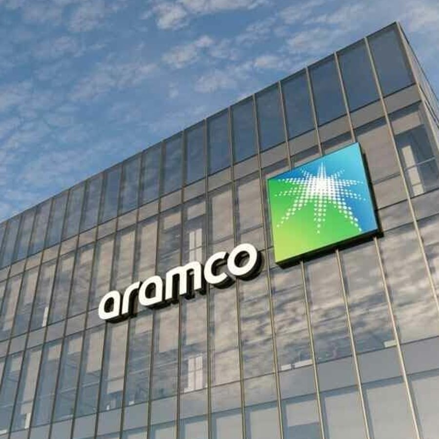 Glass building with the "aramco" logo and a green and blue star emblem on the facade, with the sky and clouds reflected in the windows.