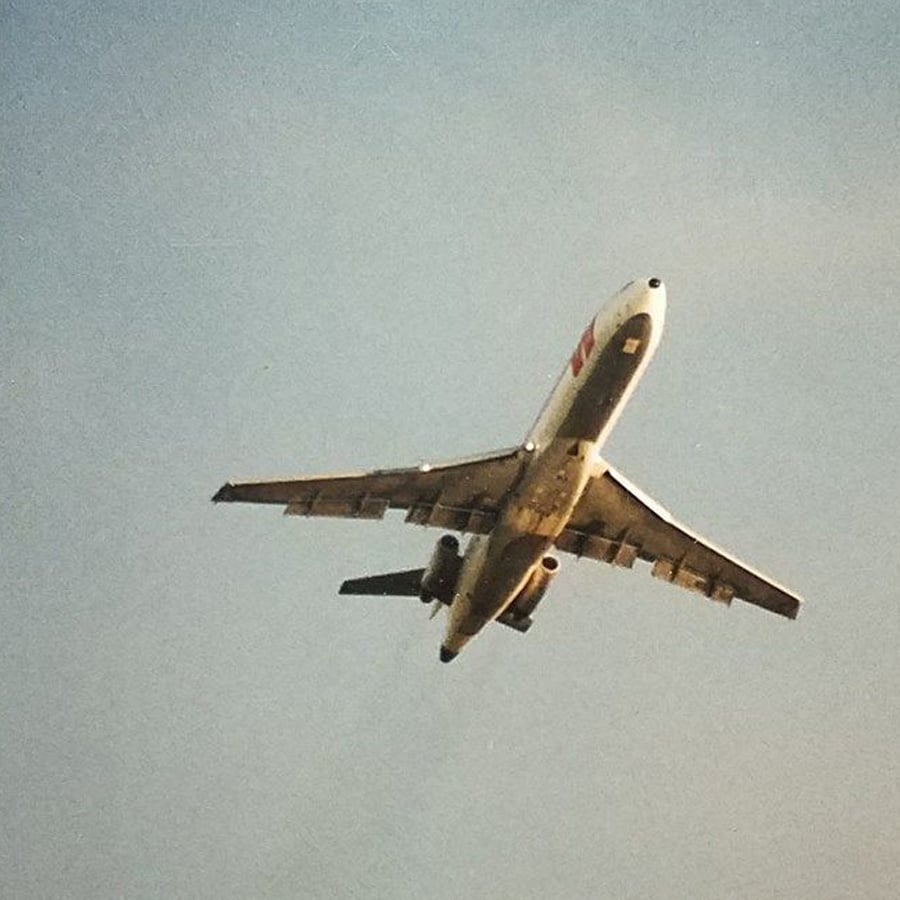 A commercial airplane is flying overhead against a clear blue sky. The underside of the aircraft is visible, showing its wings, engines, and landing gear doors.